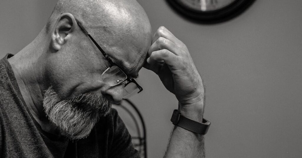 Black and white portrait of a thoughtful bald man indoors, capturing a moment of reflection with a wall clock in the background.