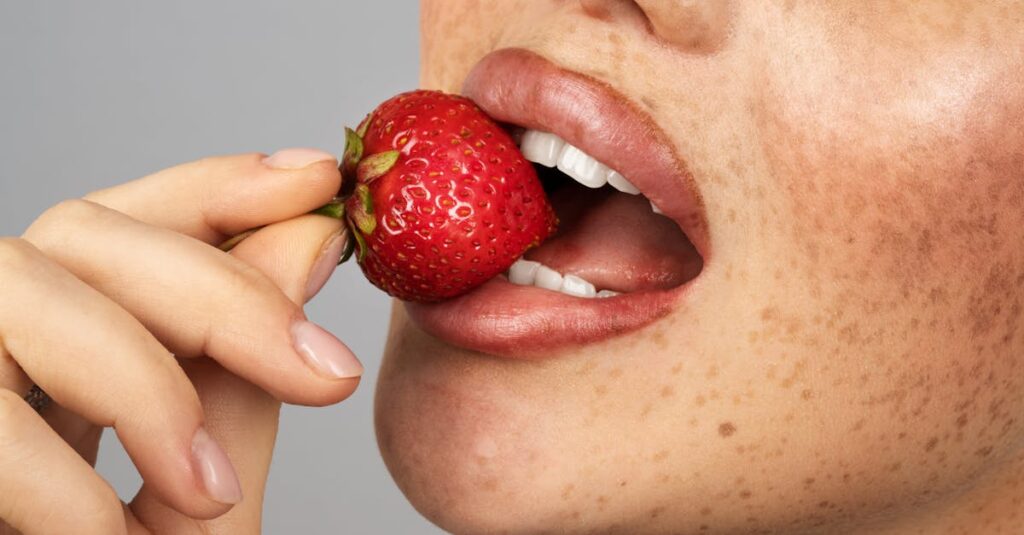 An intimate close-up of a woman enjoying a ripe strawberry with freckles visible on her face.