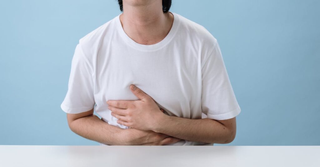 An adult man in a white shirt holds his stomach, indicating pain and discomfort, with a light blue background.
