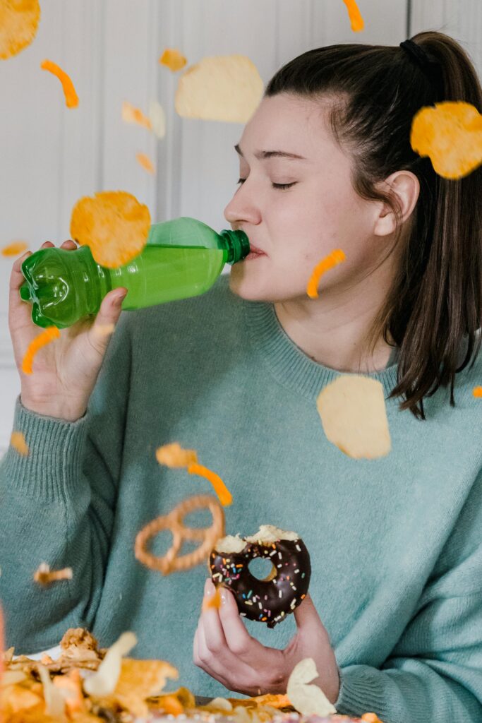 Delighted female with closed eyes showered by chips and crisps drinking fresh cold soda and eating yummy donut near heap of yellow snacks
