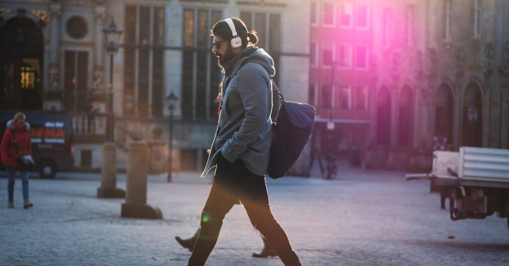 Man with headphones walks down a sunlit urban street, casting a shadow on the cobblestones.
