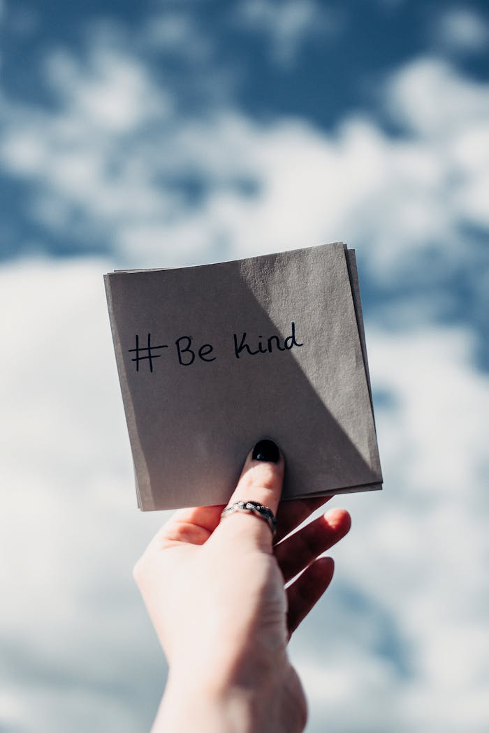 Home A hand holding a note with '#Be kind' inscribed, set against a bright sky.