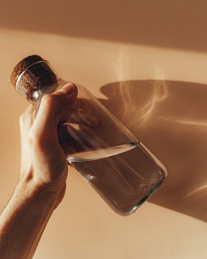 Home Crop anonymous male holding transparent glass bottle of water in hand against beige wall with shades during sunny day