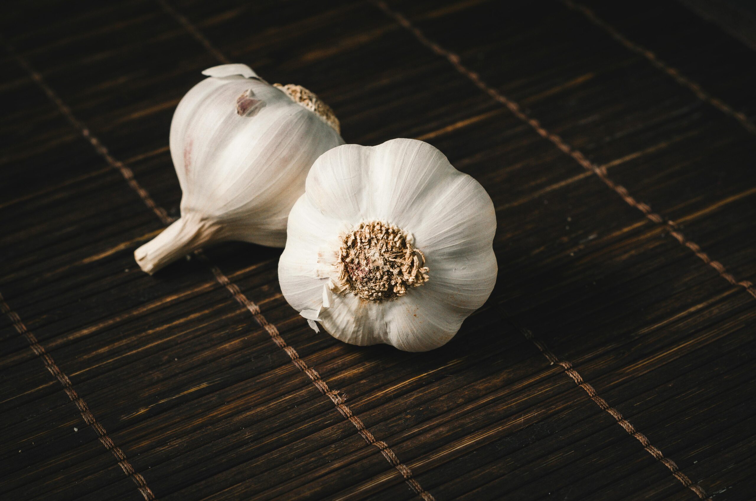 A Clove a Day Keeps the Pills Away: How Not To Die Series - Post # 2 Two fresh garlic bulbs on a dark wooden mat, highlighting aromatic culinary ingredients.